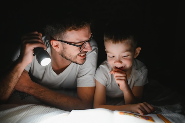 Famiglia felice che legge la favola della buonanotte sotto la coperta la sera Padre e figlio trascorrono del tempo insieme la festa del papà
