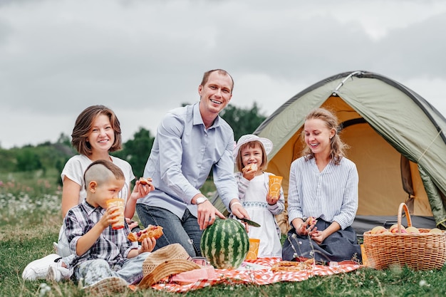 Famiglia felice che mangia anguria al picnic nel prato vicino alla tenda Famiglia godendo le vacanze in campeggio in campagna