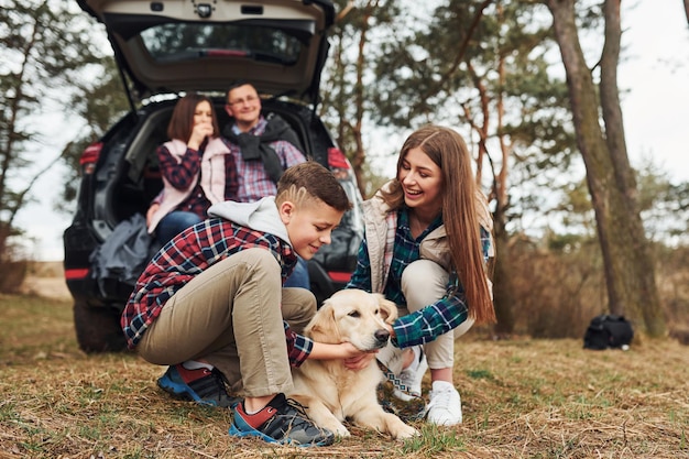 Famiglia felice che si siede e si diverte con il proprio cane vicino a un'auto moderna all'aperto nella foresta