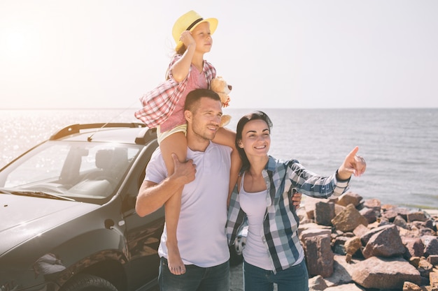 Famiglia felice che sta vicino ad un'automobile sulla spiaggia. Famiglia felice in viaggio sulla strada. Papà, mamma e figlia viaggiano in riva al mare o sull'oceano o sul fiume. Giro estivo in automobile