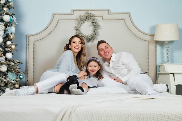 Famiglia felice con i regali di Natale sul pavimento a casa il giorno di buon Natale e felice anno nuovo. Famiglia con cane che si diverte vicino all'albero di Natale in camera da letto sul letto. I genitori adorabili danno il cane alla figlia. Copia spazio