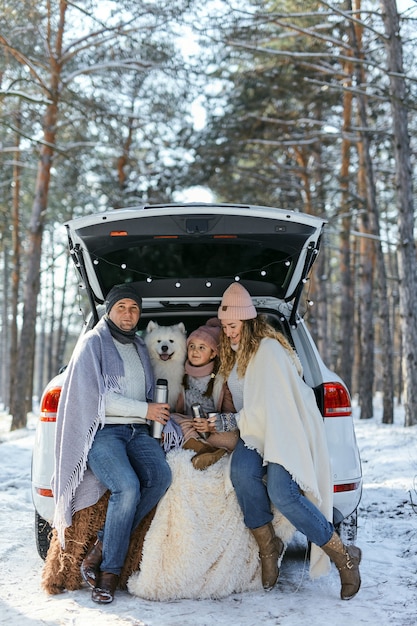 Famiglia felice con il cane in vacanza durante le vacanze invernali vicino alla strada. Vestito con abiti pesanti, seduto sul bagagliaio di un'auto e bevendo tè da un thermos. spazio per il testo. Vacanze invernali