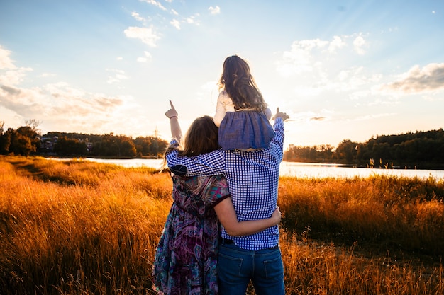 Famiglia felice con una piccola figlia in un campo in natura, guardando avanti, vista dal retro, sotto i raggi del tramonto.