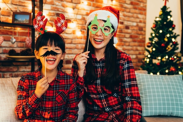 famiglia felice divertendosi a giocare con la decorazione di carta sul divano con albero di natale decorato sullo sfondo. giovane mamma con cappello da babbo natale e occhiali di natale. ragazzino che gioca con i baffi divertenti con le renne.