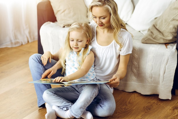 Famiglia felice. Giovane madre bionda che legge un libro a sua figlia carina mentre era seduto al pavimento di legno in una stanza soleggiata. Concetto di maternità.