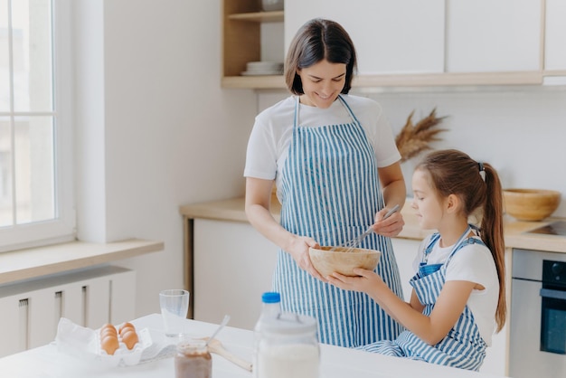 Famiglia felice in cucina. La bella donna e sua figlia preparano insieme la panetteria, indossano grembiuli, amano cucinare insieme, si godono l'atmosfera domestica, si divertono al chiuso. Bambini, maternità, concetto di cottura