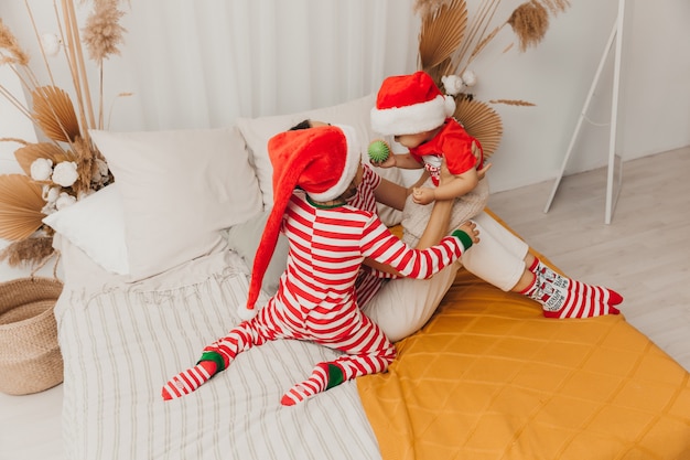 Famiglia felice, madre e bambini in pigiama e cappelli natalizi che si divertono a letto. Natale
