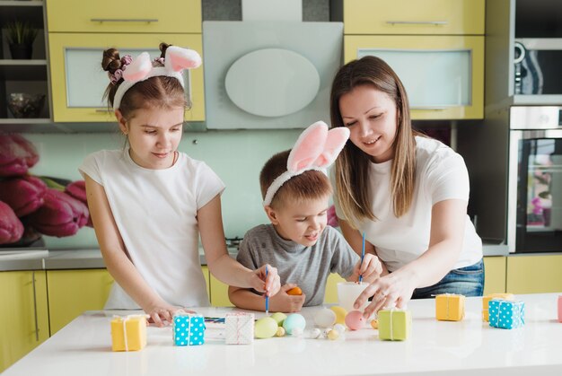 Famiglia felice: madre figlia e figlio con orecchie da coniglio si stanno preparando per le vacanze, colorando le uova nell'accogliente cucina di casa. preparativi per le vacanze di Pasqua