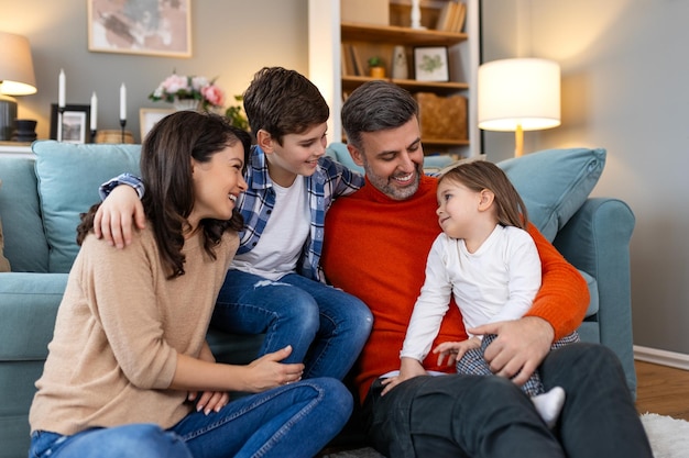 Famiglia felice seduta sul divano a ridere insieme genitori allegri che giocano con i loro figli a casa il padre face il solletico alla figlia mentre la madre e il fratello sorridono