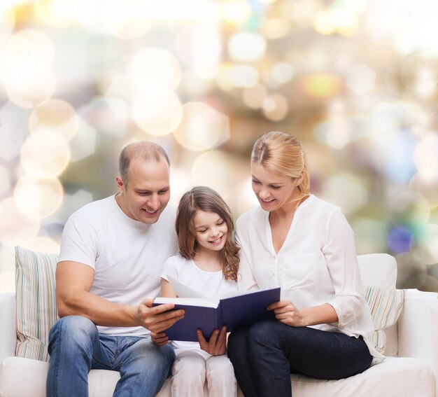 famiglia, infanzia, vacanze e persone - madre sorridente, padre e bambina che legge il libro su sfondo di luci