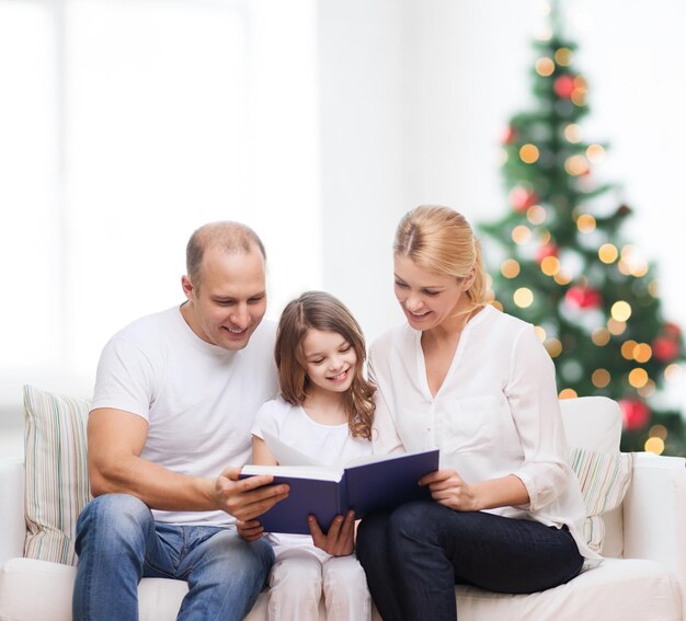 famiglia, infanzia, vacanze e persone - madre sorridente, padre e bambina che legge un libro sul soggiorno e sullo sfondo dell'albero di Natale