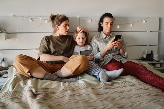 Famiglia lesbica che riposa sul letto in camera da letto con il figlio piccolo e usa i gadget