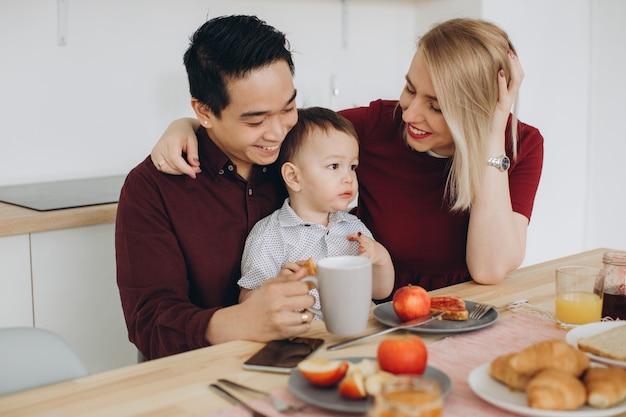 Famiglia multiculturale felice. Papà asiatico e sua moglie bionda caucasica fanno colazione con il loro bellissimo figlio in cucina.