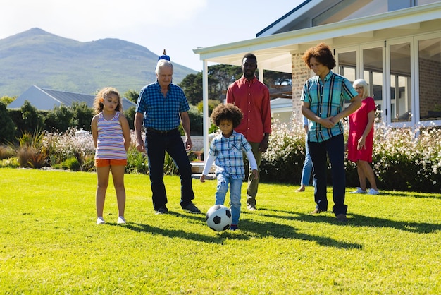 Famiglia multirazziale multigenerazionale che guarda il ragazzo che gioca a calcio sul campo erboso in cortile in estate