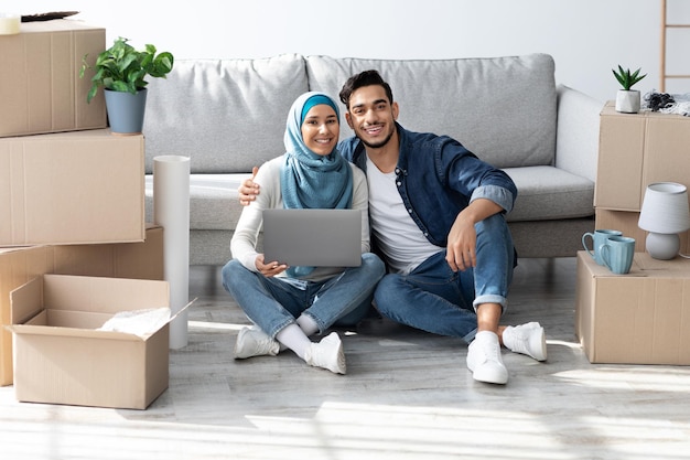 Famiglia musulmana allegra che si siede sul pavimento con il computer portatile fra le scatole