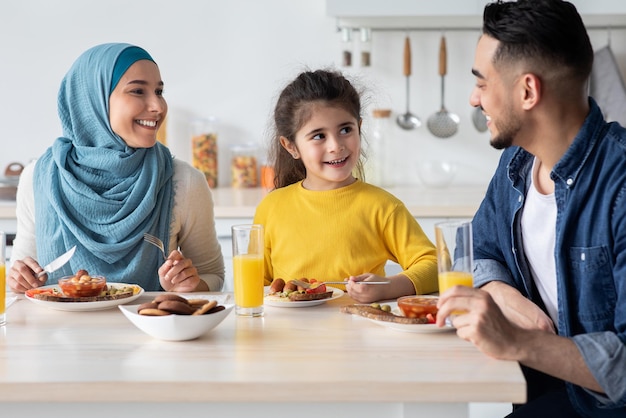 Famiglia musulmana moderna allegra con la piccola figlia che mangia la colazione insieme in cucina, madre mediorientale sorridente, padre e bambino di sesso femminile che godono del cibo saporito e che bevono il succo d'arancia