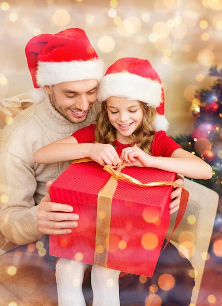 famiglia, natale, natale, inverno, felicità e concetto di persone - padre e figlia sorridenti in cappelli di Babbo Natale che aprono la confezione regalo