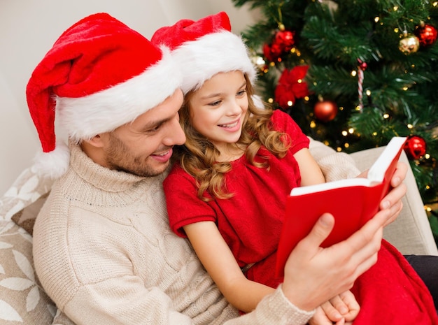 famiglia, natale, natale, inverno, felicità e concetto di persone - padre e figlia sorridenti nel libro di lettura dei cappelli di santa helper