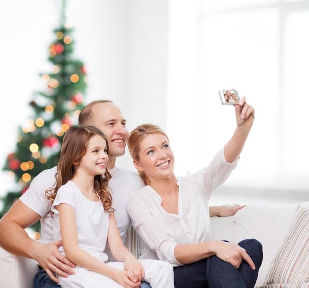 famiglia, vacanze, tecnologia e persone - madre, padre e bambina sorridenti che fanno selfie con la fotocamera sul soggiorno e sullo sfondo dell'albero di Natale