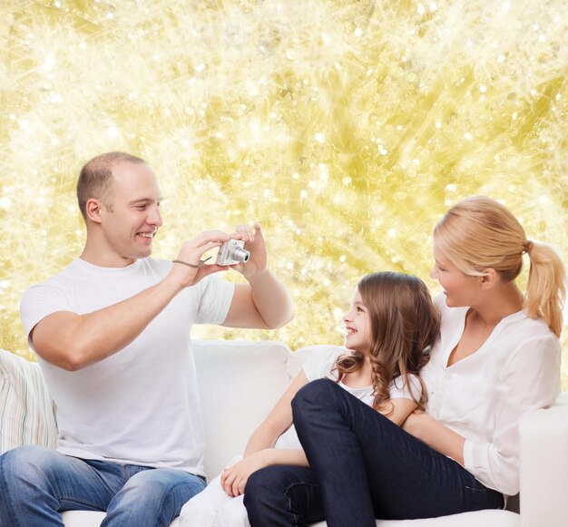 famiglia, vacanze, tecnologia e persone - madre, padre e bambina sorridenti con la fotocamera su sfondo di luci gialle