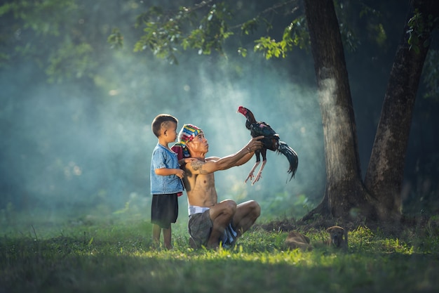Famiglie, padre e figlio asiatici del contadino con il gallo di mattina ad area locale, Tailandia