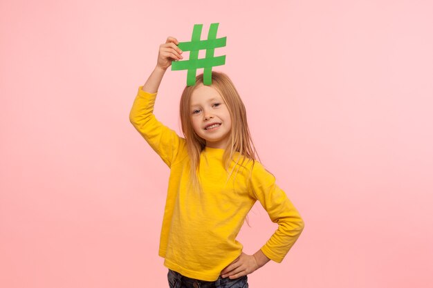 Famoso blog per bambini. Ritratto di una fantastica bambina carina che tiene il simbolo dell'hashtag sulla testa e guarda la telecamera con un sorriso a trentadue denti, mostrando il segno di hashtag. studio al coperto isolato su sfondo rosa