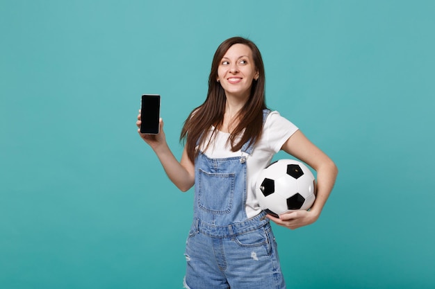 Fan di calcio da ragazza sognante che tiene pallone da calcio, telefono cellulare con schermo vuoto vuoto isolato su sfondo blu turchese. Emozioni della gente, concetto di stile di vita di svago della famiglia di sport. Mock up spazio di copia.