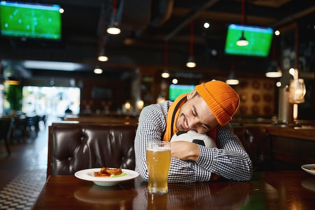 Fan di calcio divertente del giovane che dorme sulla palla mentre riposa nella barra di sport. Ragazzo annoiato che aspetta che gli amici guardino la partita al pub