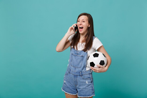 Fan di calcio eccitato ragazza allegra con pallone da calcio che parla sul telefono cellulare conducendo una piacevole conversazione isolata su sfondo blu turchese. Emozioni della gente, concetto di svago della famiglia di sport.