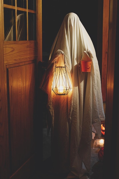 Fantasma spaventoso con lanterna incandescente e secchio di caramelle di Halloween alle porte nella spettrale notte atmosferica Felice Halloween persona vestita da fantasma dolcetto o scherzetto all'ingresso di casa al buio