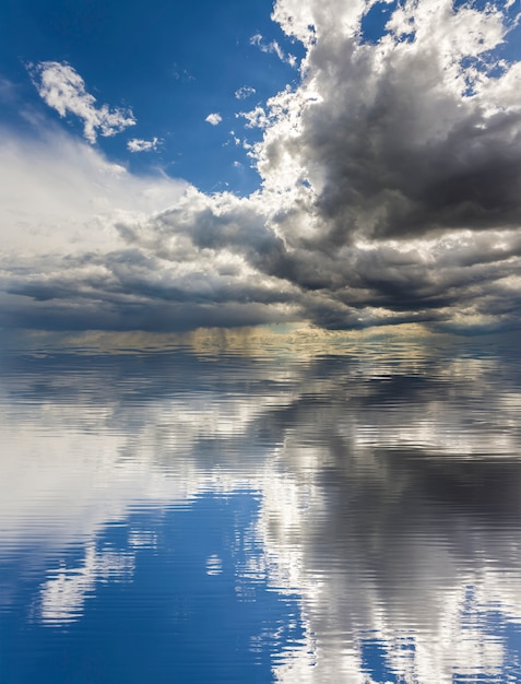 Fantastica vista di luminose nuvole gonfie bianche e scure illuminate dal sole contro il cielo blu profondo che si riflettono nell'acqua. Bellezza e potenza della natura, problemi ambientali, ecologia e concetto di cambiamento climatico.