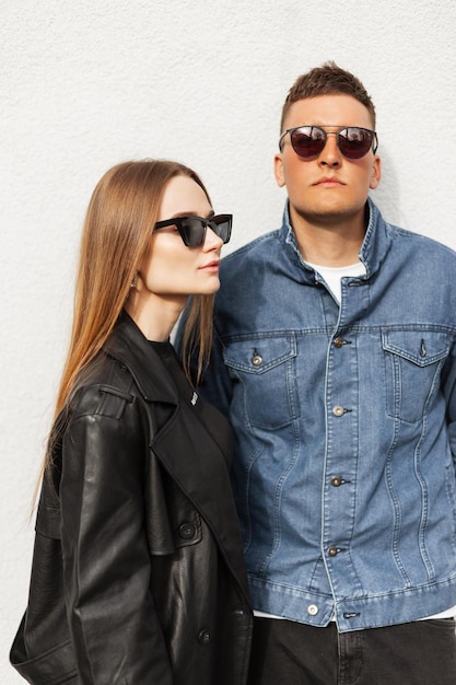 Fantastico ritratto di strada di una bella giovane coppia con occhiali da sole vintage che indossa abiti casual alla moda con una giacca di jeans e un appendiabiti in pelle vicino al muro Ragazza alla moda e uomo alla moda