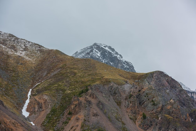 Fantastico scenario con la cima di una montagna innevata con rocce affilate sopra una grande montagna coperta di muschio con alberi sotto il cielo nuvoloso Paesaggio suggestivo con alta collina di muschio e cima di una montagna innevata con tempo variabile