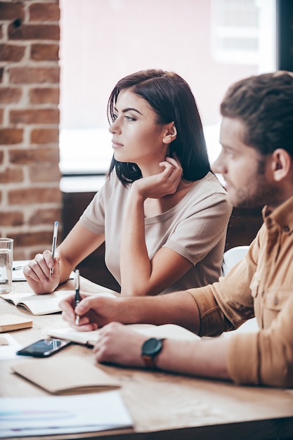 Fare alcune note importanti. Vista laterale della giovane e bella donna pensierosa che tiene la mano nei capelli mentre è seduta con i suoi colleghi al tavolo