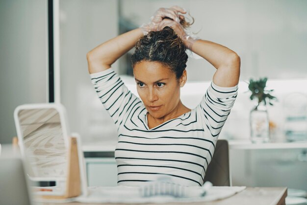 Fare bellezza cura acconciatura persone concetto stile di vita attività per il tempo libero al coperto a casa Una bella giovane donna adulta che si fa acconciatura guardandosi allo specchio in cucina Industria della moda capelli