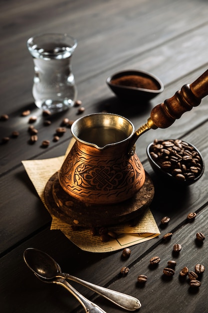 Fare il caffè a cezve. Fagioli, caffè macinato, cezve, acqua in un bicchiere, cucchiaio. Fondale in legno.