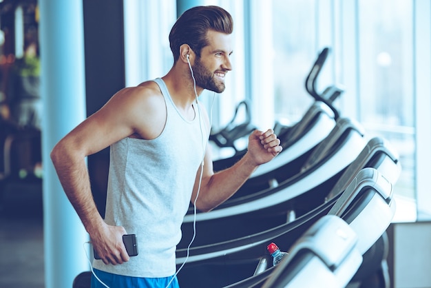 Fare jogging con piacere. Vista laterale del giovane bell'uomo in cuffia che guarda lontano con un sorriso mentre corri sul tapis roulant in palestra