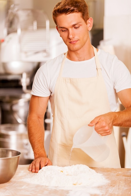 Fare la pasta per la pasticceria. Giovane fornaio maschio sicuro che versa acqua sulla farina