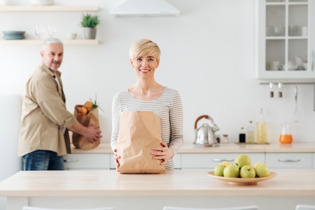 Fare la spesa in famiglia online. Ritorno a casa dopo il mercato o la spesa. Felice donna europea senior e maschio con pacchetti ecologici con verdure dopo il supermercato all'interno della cucina, spazio vuoto