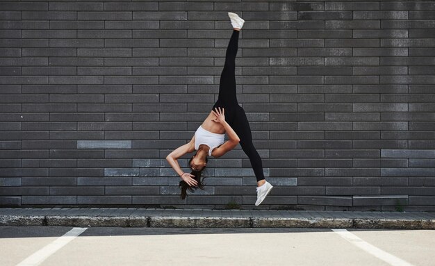 Fare parkour pazzo e acrobazie atletiche Giovane bruna sportiva con forma di corpo sottile contro il muro di mattoni in città di giorno