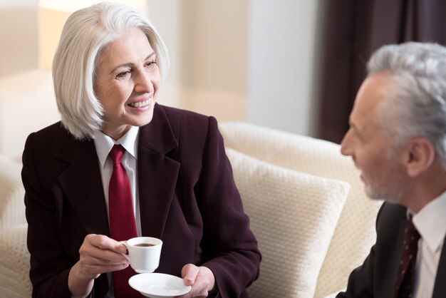 Fare una pausa. Allegra imprenditrice invecchiata felice seduta in hotel mentre discute il progetto con il suo collega e beve caffè