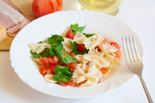 Farfalle di pasta con pomodori ed erbe aromatiche, forchetta, pancetta, formaggio, pomodoro e olio in una brocca su sfondo bianco