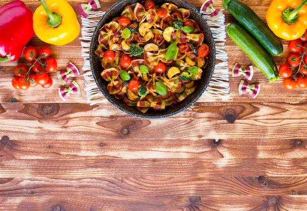 Farfalle di pasta italiana in salsa di pomodoro e vari tipi di verdure su un fondo di legno, spazio libero per il testo.