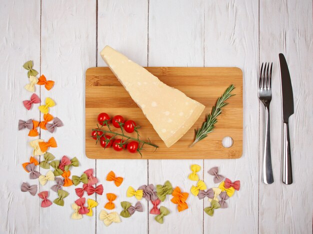 Farfalle di pasta italiana secca con pomodori, formaggio, rosmarino, forchetta e coltello su fondo in legno chiaro. Con copia spazio