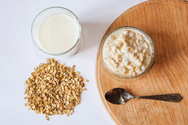 Farina d'avena in una ciotola con un bicchiere di latte, un cucchiaio e farina d'avena su una tavola di legno. Vista dall'alto.