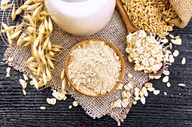 Farina d'avena in una ciotola, latte in brocca, farina d'avena in cucchiaio su tela, grano in borsa, gambi di avena su tavola di legno sfondo dall'alto