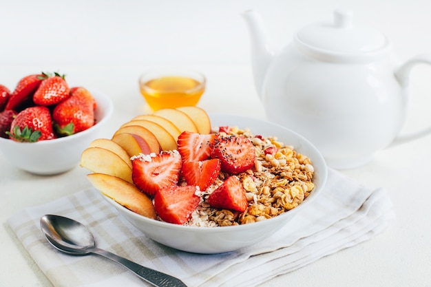 Farina d'avena porridge di colazione con cereali muesli fragole pesche miele in un piatto bianco sullo sfondo di una teiera bianca e ciotole con miele su un piatto bianco