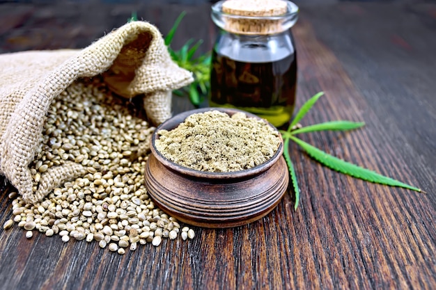 Farina di canapa in una ciotola di argilla, il grano nel sacchetto e sul tavolo, l'olio in un barattolo di vetro, foglie e gambi di cannabis su uno sfondo di assi di legno