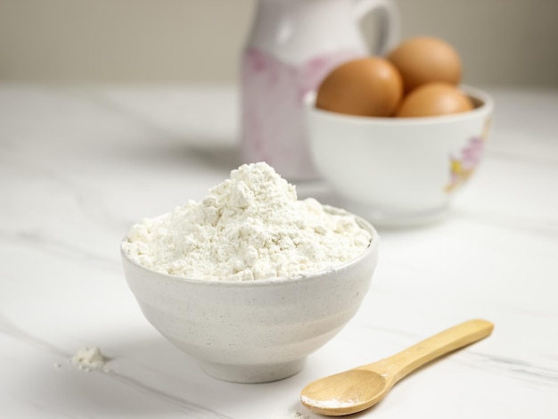 Farina di frumento in una ciotola bianca con uova e latte come sfondo. Concetto di preparazione al forno.