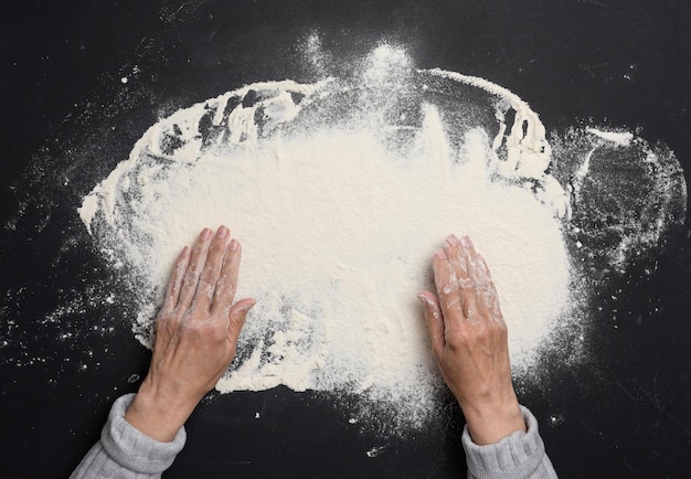 Farina di grano bianco setacciata su un tavolo nero e due mani femminili in vista dall'alto che cucinano a casa